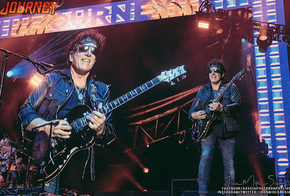Neal Schon Journey Founder plays Guitar against a Journey banner
